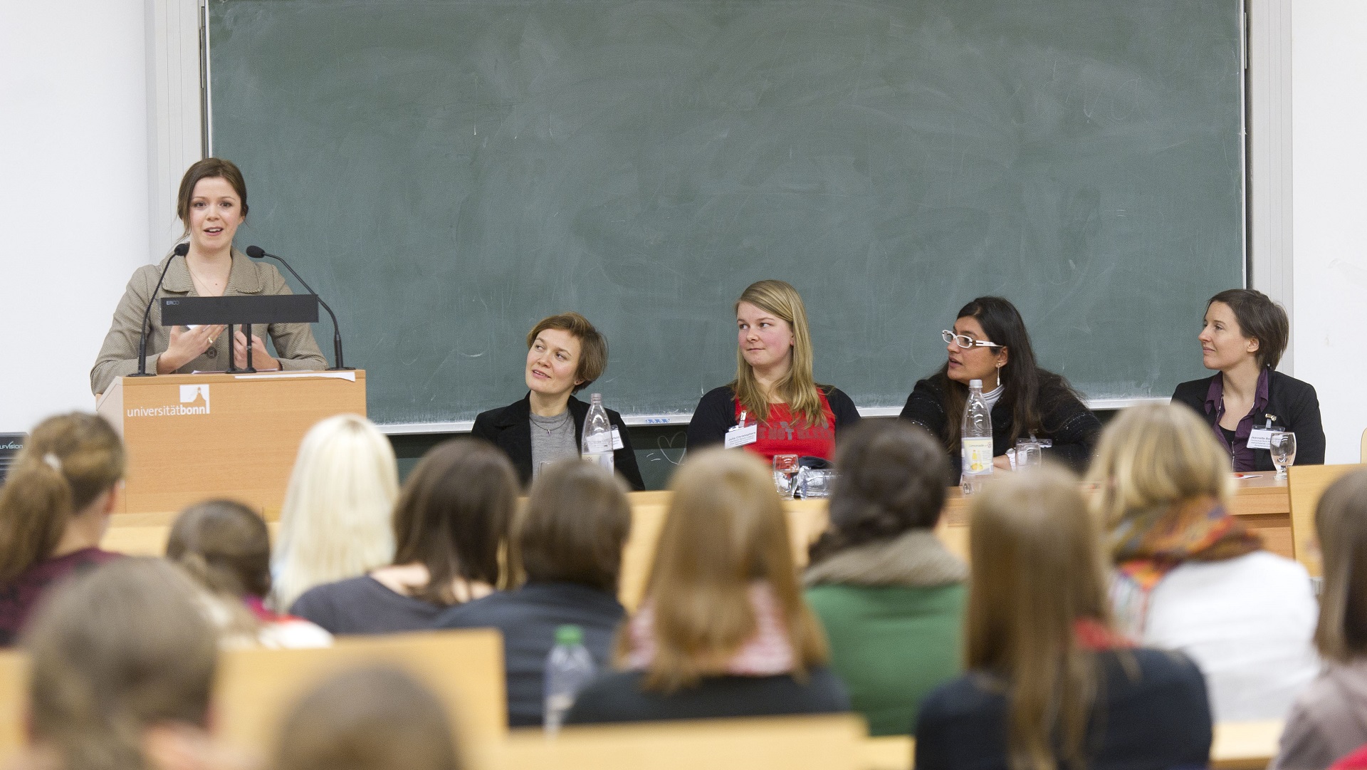 Vier weibliche Personen auf einem Podium im Hörsaal, eine der Personen spricht zu den Zuschauern.